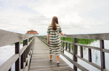 Lady Strolling on a Wooden Boardwalk Towards a Pavilion in Bueng Bua Wetland. Woman in a striped...