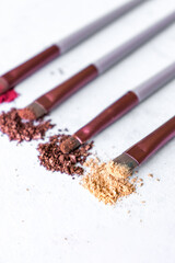 A high resolution close up shot of four makeup brushes arranged in a row, each dipped in different shades of crushed eyeshadow pigment pink, bronze, brown, and beige. The brushes lie flat on a white