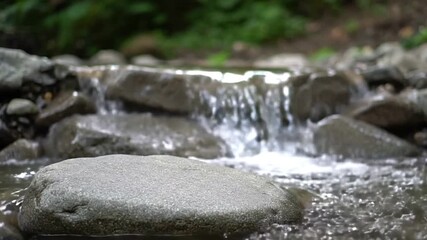 A stream flows over rocks with a smooth round stone in the foreground Green foliage is visible in the blurred background
