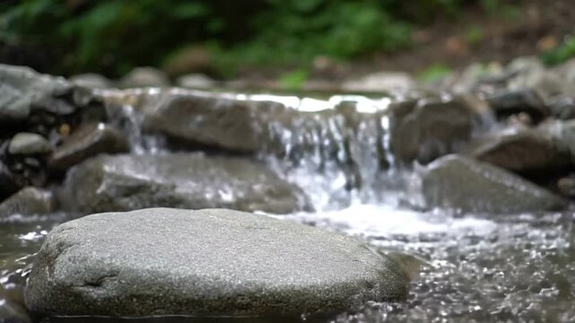 A stream flows over rocks with a smooth round stone in the foreground Green foliage is visible in the blurred background