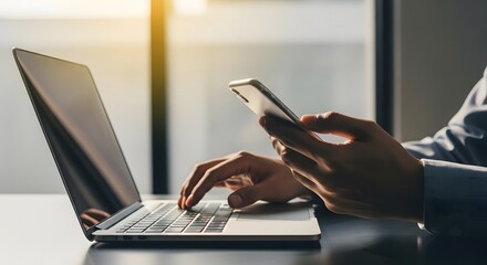 Fototapeta premium A person's hands typing on a laptop keyboard while holding a smartphone, illustrating modern digital work and communication in a professional setting