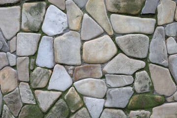 A detailed texture of a wall paved with uneven stones of various shapes and shades of gray and beige. Green moss is visible between the stones, giving the surface a natural and antique look.