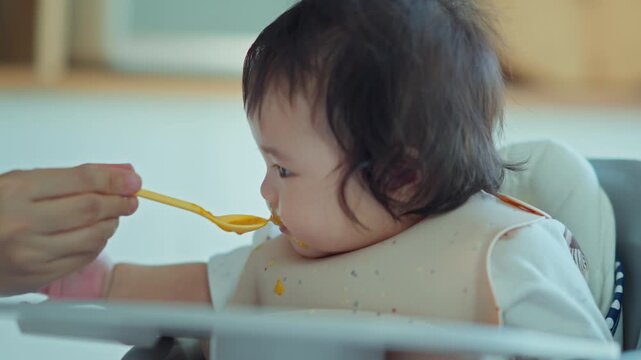 Close-up of happy Asian baby girl eating her first solid food with pumpkin puree from spoon with mom at home &ndash; adorable infant smiling and looking at camera, baby weaning, parenting and family moment