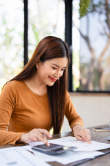  Happy young asian entrepreneur woman in casual counting profit, on calculator at laptop computer,...