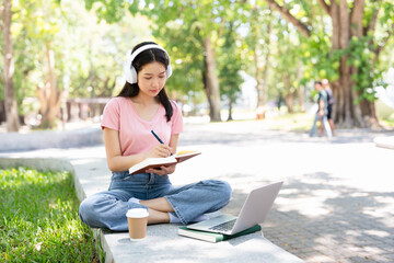 Smiling young asian woman in wireless headphones sitting outdoor working on laptop computer and...