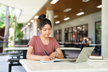 Focused asian teen girl student writing notes in notebook distance learning studying online makes...