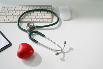 Stethoscope, red heart model, and keyboard on white medical desk. Healthcare and cardiology concept