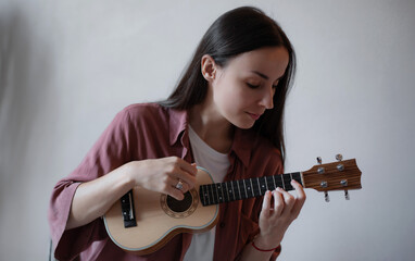 Young woman playing a ukulele at home with focus and calm expression, wearing casual clothes and...