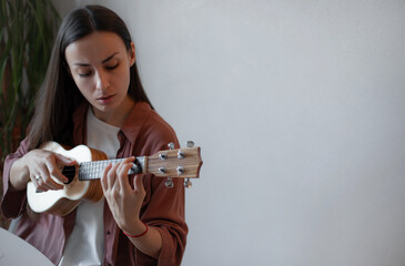 Young woman with long brown hair intently playing ukulele at home, wearing a rust shirt and white...