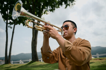 Man in sunglasses playing trumpet sitting on green grass with scenic mountain backdrop
