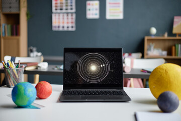 Laptop displaying solar system diagram standing on desk in classroom with colorful planet models in foreground, educational materials and charts visible in background