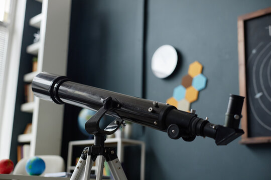 Telescope standing on tripod in classroom with blurred educational objects in background, showing astronomy learning environment for students interested in science and space exploration