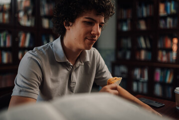 Student eating sandwich and studying in library with bookshelves