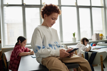 Caucasian boy sitting on desk using smartphone while two other children working in background,...