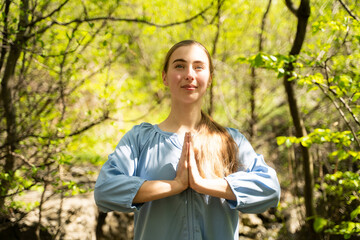 Calm young woman meditating hands in namaste gesture in green forest with sunlight, breathing fresh air outdoors, enjoying the peaceful moment in summer day, healthy lifestyle, front view portrait
