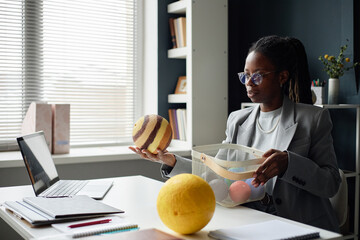 Young adult Black woman sitting at desk with laptop holding planet model in hand, while explaining...