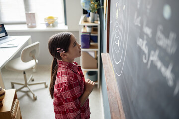Caucasian girl child standing at chalkboard studying solar system diagram in classroom setting,...