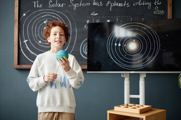 Caucasian boy child standing in classroom holding model of Earth and smiling, presenting solar system project with digital display and chalkboard diagram in background