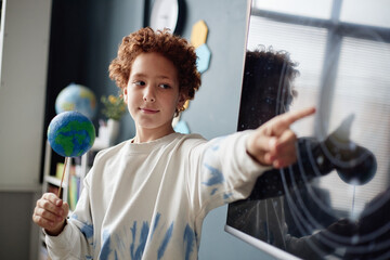 Caucasian boy child holding model of Earth, standing in classroom and pointing at solar system...