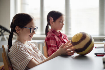 Caucasian girl child with glasses sitting at desk smiling and touching planet model, while another...