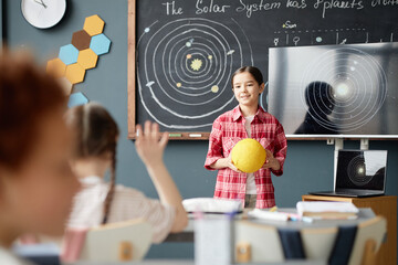 Caucasian girl child standing in classroom holding yellow model of Sun, presenting in front of...