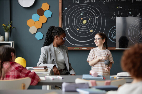 Black woman teacher interacting with Caucasian girl student holding model planet in classroom, other children sitting at desks, chalkboard showing solar system diagram in background