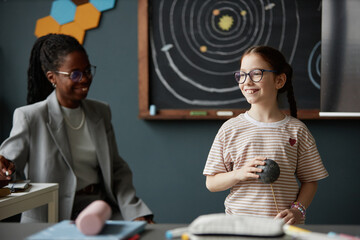 Caucasian girl smiling while holding model planet in classroom, Black woman teacher sitting nearby,...
