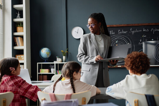 Black woman teacher holding file folder standing in front of classroom engaging with group of diverse children sitting at desks, listening attentively during science lesson about solar system