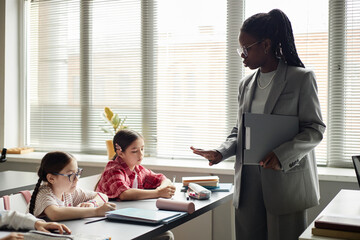 Black woman teacher standing and gesturing while two Caucasian girls sitting at desk writing in...