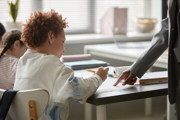 Caucasian boy sitting at desk writing while adult Black woman standing beside him pointing at...