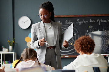 Black woman teacher standing in classroom holding clipboard and pen instructing diverse group of...
