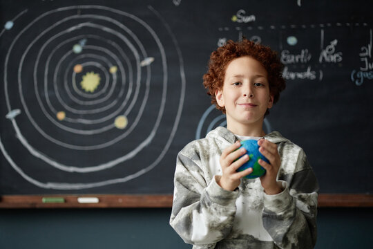 Portrait of Caucasian boy child holding model of planet while standing in front of chalkboard with solar system diagram, looking into camera with positive expression in classroom