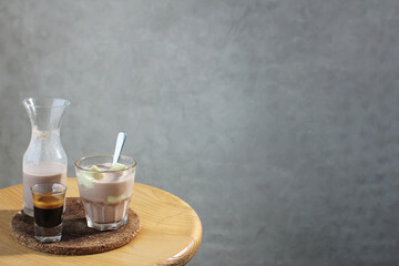 
A creative coffee preparation features a glass of chocolate milk with ice, a shot of espresso, and a small carafe of coffee-infused milk on a rustic wooden table against a grey concrete wall