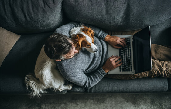 A top view of a man wearing yellow working on his laptop while sitting on the sofa with a dog lying next to him - Powered by Adobe