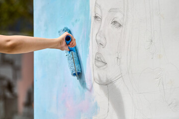 Hand applies soft blue paint over pencil sketch of young female face on white wall. Natural outdoor...