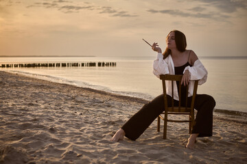 Young woman, styled as painter, sits contemplatively on chair at beach during sunset. Serene...