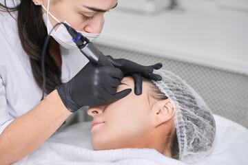 Young woman receives eyebrow permanent makeup in serene spa. Beauty technician, wearing gloves,...