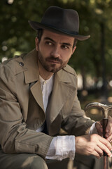 Man in vintage attire sits on a park bench under trees holding a cane. Concept of promotional...