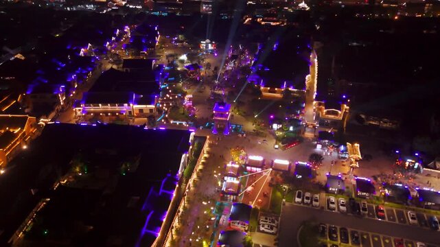 Drone video shows Pingyao Ancient City at night, aglow with colorful lights on traditional buildings. A lively scene unfolds below, full of activity beneath a starry sky.