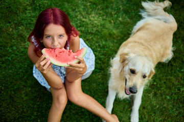 Teen girl enjoying watermelon while sitting on grass with a dog nearby during a sunny day in the backyard