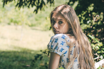 A young woman with long hair sits among greenery, dressed in a floral pattern, looking back with a...