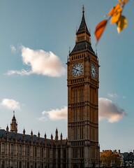 Iconic big ben clock tower bathed in golden sunlight against a clear blue sky with wispy clouds and autumn leaves