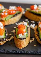 close up stock photo of greek-style crostini topped with olive paste, cucumber slices, cherry tomatoes, and feta cheese