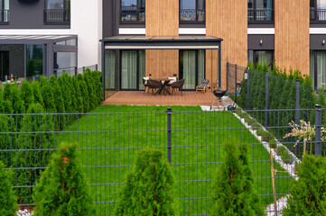 modern backyard patio with seating area and grill. green lawn surrounded by trimmed hedges and metal fencing enhances cozy outdoor space. ideal for relaxation, small gatherings, family time.