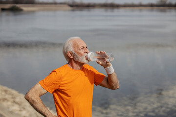 Thirsty tired senior sportsman is drinking water on riverside.