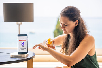 Smartphone with high UV alert in focus, while woman in background applies sunscreen to her skin....