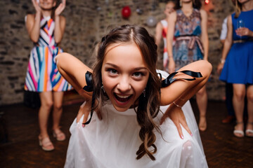 Fototapeta premium Close-up of an excited young girl leaning forward, looking directly at the camera at a vibrant party. Blurred figures and decorations are in the background, capturing a moment of pure joy