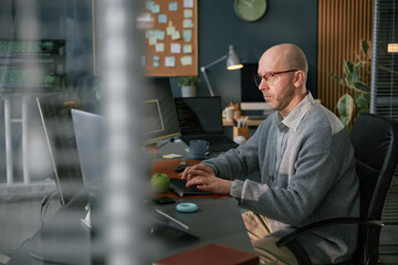 Fototapeta premium Middle aged Caucasian man working at computer in modern office, typing on keyboard and focusing on screen, surrounded by technology devices and office supplies
