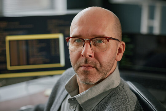 Portrait of middle aged Caucasian man wearing glasses looking at camera while sitting at desk with computer monitors displaying programming code in modern office setting - Powered by Adobe