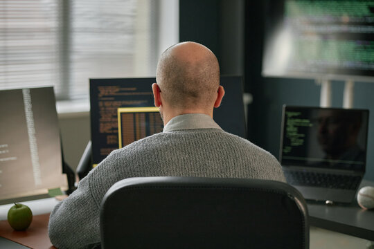 Middle aged Caucasian man sitting at desk working on multiple computer monitors with code displayed, back of head visible, office environment with window blinds and electronic devices